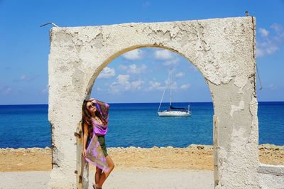Woman leaning on old arch structure by sea against sky