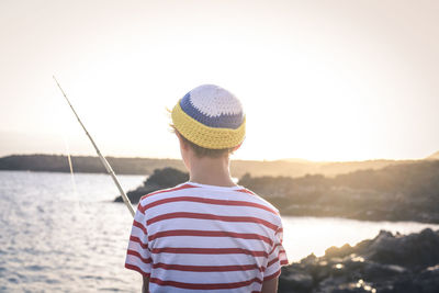 Rear view of boy standing with fishing rod by sea