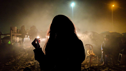 Rear view of silhouette woman standing by illuminated lights at night