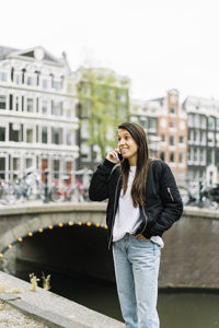 Happy young ethnic female tourist with long dark hair in trendy outfit speaking on mobile phone while standing against typical buildings in amsterdam near canal and bridge