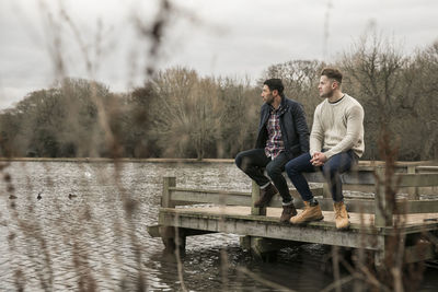 Friends looking away while sitting on pier over lake in epping forest