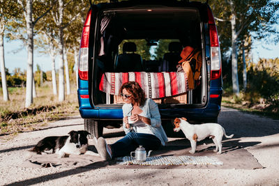Woman holding drink while sitting with dogs by camper trailer