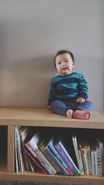 Cute baby boy sitting on bookshelf against wall at home