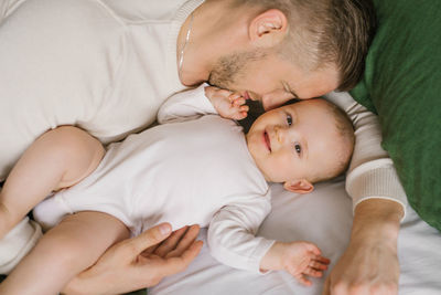 A handsome young dad lies in bed in the bedroom and hugs his loving laughing six-month-old boy.