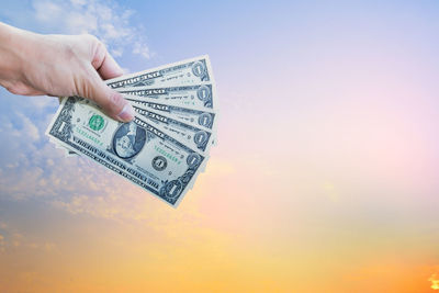 Cropped hand holding paper currency against sky during sunset
