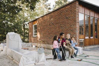 Parents with children eating food while resting on concrete block against house