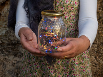 Midsection of woman holding string lights in jar against stone wall