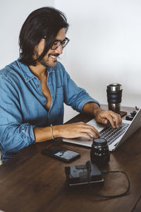 Smiling man using laptop computer on table while sitting against wall at home