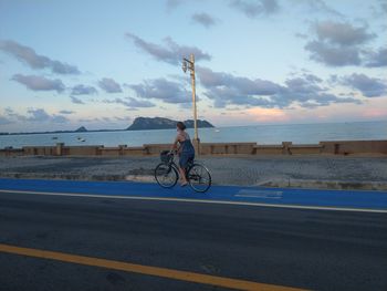 Man riding bicycle on road by sea against sky