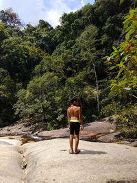 Rear view of shirtless boy standing on rock against trees in forest