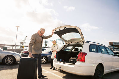 Senior man loading luggage in car trunk with woman standing in parking lot against sky