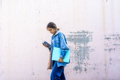 Teenage girl walking with book and bottle by wall on sunny day