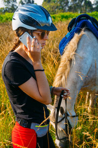 Young woman using phone while standing with horse against sky