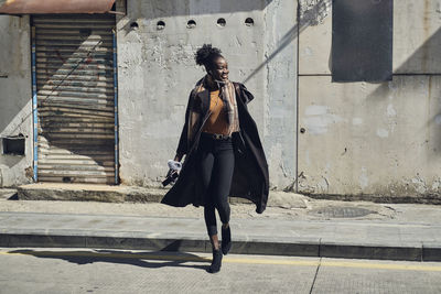 Young glad african american female in trendy clothes with earphones walking on asphalt roadway while looking away in city