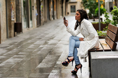 Full length of young woman using mobile phone while sitting on bench in city