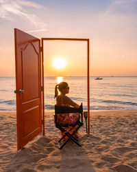 Rear view of woman sitting on beach against sky during sunset