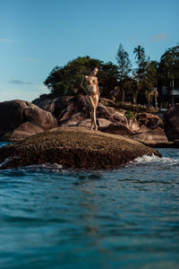 A pregnant girl in gold glitter stands on a coral stone in the middle of the ocean