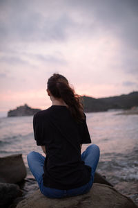 Rear view of woman sitting on rock at beach during sunset