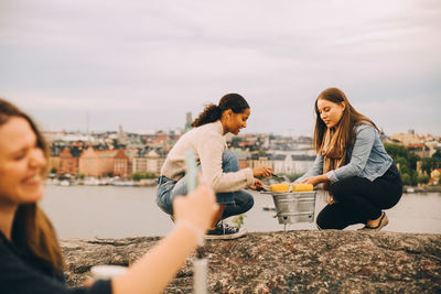 Female friends roasting sweetcorn on rock formation by lake during picnic