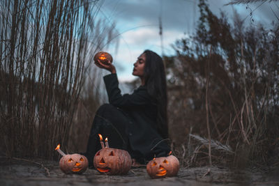 Full length of woman sitting by pumpkin on tree during halloween