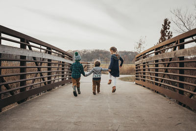 Three siblings holding hands running on bridge away from camera