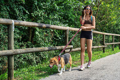 A happy girl in sunglasses and sportswear on the phone walks with her beagle dog on a walking track