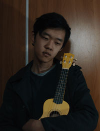 Teenage boy holding ukulele against wooden door