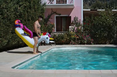 Side view of shirtless man standing with unicorn inflatable ring at poolside