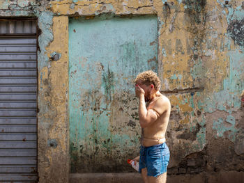 Full length of shirtless man standing against wall with hand to face.