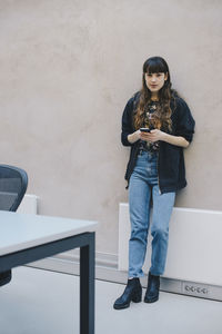 Full length portrait of female computer programmer holding smart phone while standing against beige wall in office