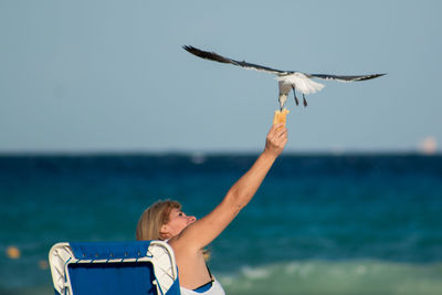 Rear view of woman feeding seagull flying over beach