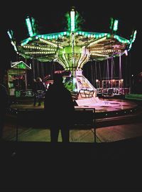 Rear view of silhouette person at illuminated amusement park against sky at night