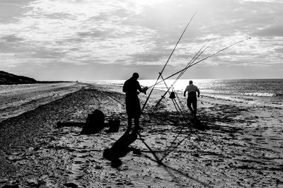Silhouette men fishing on beach against sky