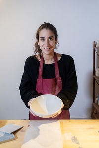 Smiling young ceramist woman showing ceramic bowl in pottery workshop