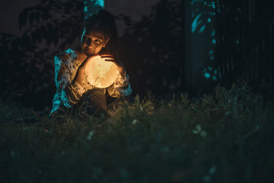 Girl holding illuminated toy sitting on field at night