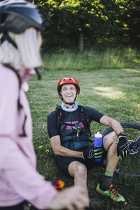 Happy male cyclist sitting on grass holding water bottle while talking to female friend