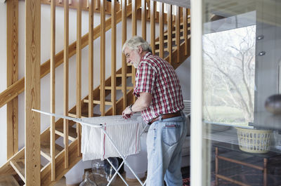 Senior man hanging clothes on drying rack while standing by wooden steps at home