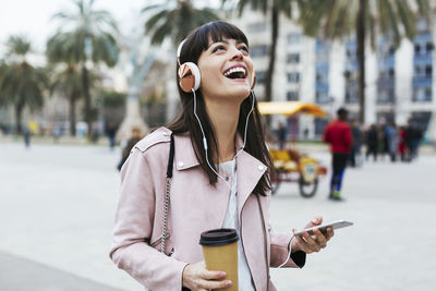 Spain, barcelona, laughing woman with coffee, cell phone and headphones in the city