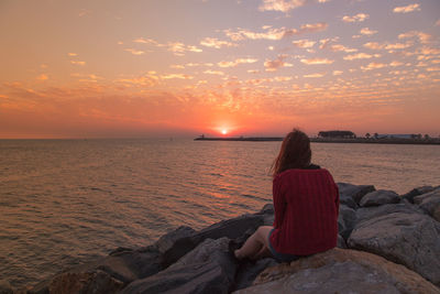 Woman sitting on rock by sea against sky during sunset