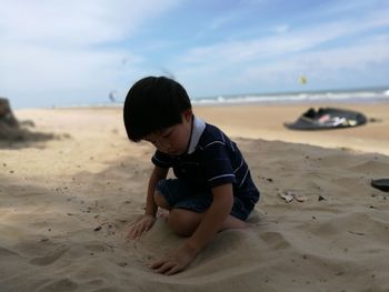 Cute boy playing with sand against sky at beach