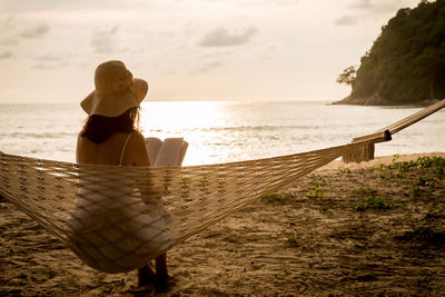 Rear view of woman reading book while sitting on hammock at beach during sunset