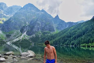 Rear view of shirtless man looking at lake against mountains