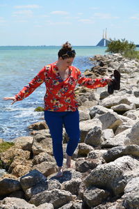 Woman holding slippers walking on rocky sea shore against sky