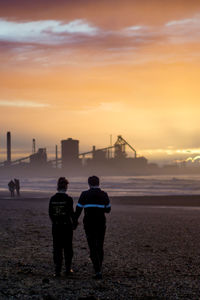 Rear view of men standing on sidewalk against sky during sunset