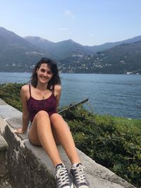 Portrait of smiling young woman sitting on retaining wall by lake