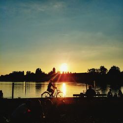 Silhouette people sitting on riverbank against sky during sunset