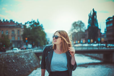 Portrait of young woman standing against sky in city