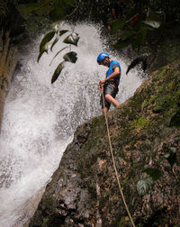 Low angle view of man tying rope while standing on cliff against waterfall