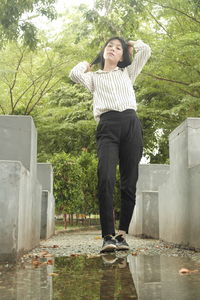 Low angle portrait of young woman standing by puddle against trees