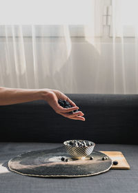 A woman's hand holding blueberries. underneath is a little bowl with more blueberries.